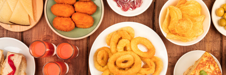 A panorama of Spanish tapas, an overhead photo of a variety of snacks. Gazpacho, squid rings, tortilla, cheese etc, shot from above on a dark rustic wooden background