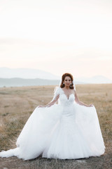 Fototapeta na wymiar Happy bride in wedding dress posing on the road in a field at sunset. Beautiful woman. Bride at wedding day walking Outdoors on nature. Beautiful and stylish bride in wedding day