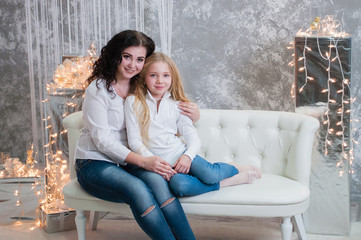 Obraz premium Two very pretty girls, sisters celebrates Christmas, new year. Girls in the New Year's interior of the room are sitting on a white sofa against the background of garlands