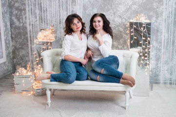 Obraz premium Two very pretty young women celebrate Christmas, the new year. Girls in the New Year's interior of the room are sitting on a white sofa against the background of garlands