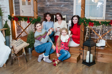 Fototapeta na wymiar Several very pretty women and a teenager girl celebrate Christmas, the new year. Girls in the New Year's interior with cups in their hands are sitting on the steps
