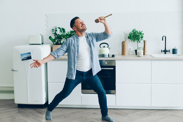 Portrait of playful middle-aged man singing in spoon in kitchen having fun standing in room alone...