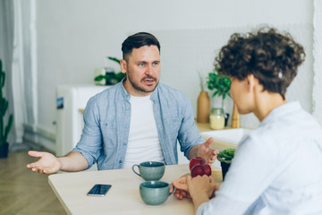 Annoyed man husband is yelling at unhappy wife in kitchen fighting gesturing sitting at table...