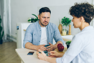 Aggressive guy husband is fighting with unhappy woman in kitchen looking with angry face sitting at table together. Conflict, problems and family concept.