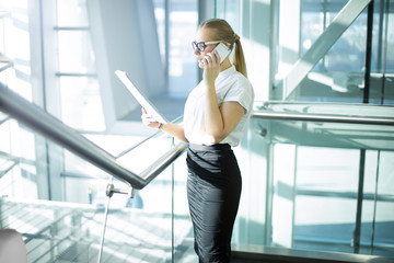 Confident female candidate in formal wear talking via mobile phone and reading summary while standing inside company. Woman in glasses business owner with paper documents in hand having cellphone talk