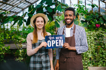 Man and woman happy business partners are holding we are open sign in greenhouse smiling looking at camera inviting people for organic food and flowers.
