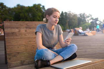 Fototapeta na wymiar Stylish hipster girl using apps on cellphone while sitting with netbook on bench in park during summer vacation. Female blogger reading article on mobile phone