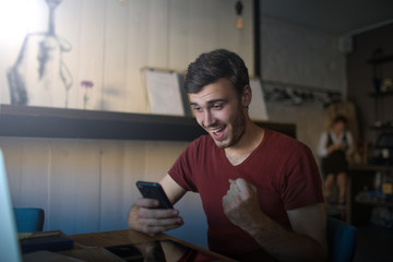 Happy smiling man freelance worker celebrating victory with winning gesture while reading on mobile phone e-mail with good news while sitting with netbook in coffee shop interior