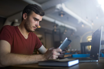 Man skilled freelance social media content writer using applications on touch pad gadget while sitting at the table with open laptop computer in modern restaurant interior. Hipster guy online ordering