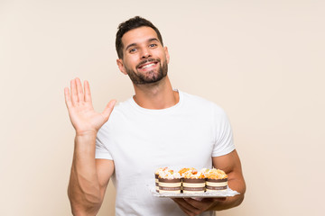 Handsome man holding muffin cake over isolated background saluting with hand with happy expression