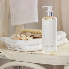 A bottle with shower gel, a body brush and a pair of bath mitts on a white chair into a bathroom 