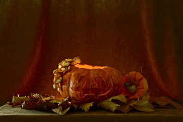 A ripe orange pumpkin, with light from a candle inside, and with a voluminous carved out silhouette of a face, with a cap lying next to it cut out from a cap with a green handle and seeds hanging from