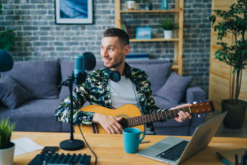 Handsome guy is playing the guitar recording audio podcast in studio using microphone and laptop sitting at table indoors alone. People and music concept.