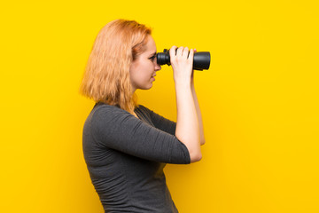 Young woman over isolated yellow background with black binoculars