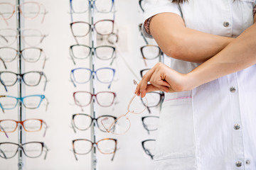 Female ophthalmologist with crossed arms in clinic. Cropped image of attractive young female doctor in ophthalmology clinic. Doctor ophthalmologist is standing near shelves with different eyeglasses.