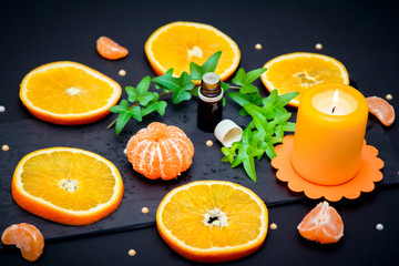 Citrus aroma oil concept – close up glass bottle with essence, ripe juicy orange and tangerine slices, candle and ivy branch with green leaves on black background.