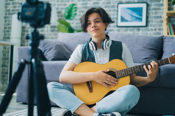 Teenage musician young boy is recording video holding guitar using modern camera in apartment....