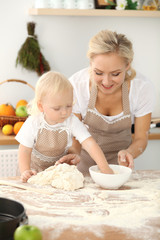 Little girl and her blonde mom in beige aprons playing and laughing while kneading the dough in kitchen. Homemade pastry for bread, pizza or bake cookies. Family fun and cooking concept