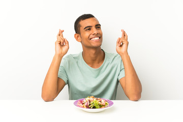 Young handsome man with salad in a table with fingers crossing