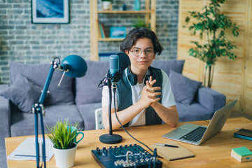 Teenager using microphone and laptop recording podcast in apartment sitting at table looking at...