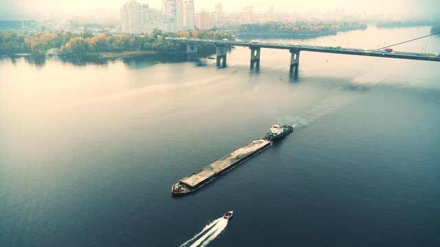 Scenic aerial cityscape of Kiev and river Dnipro at sunset. Tugboat supporting barge with sand bulk materials heading down river Dnieper. Ukrainian inland navigation and river freight cargo