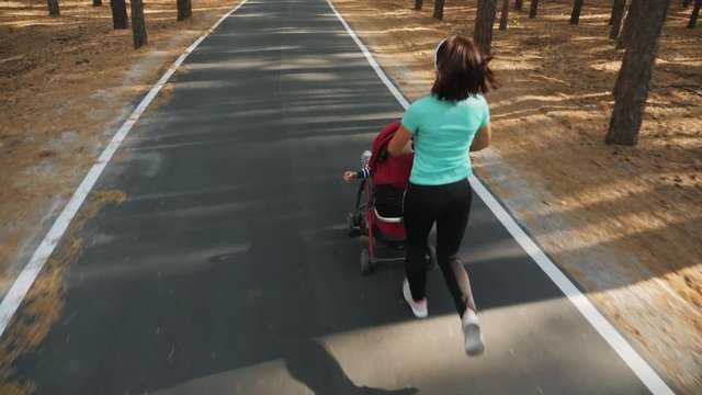 Top rear view: Active athletic woman running with a baby stroller on an asphalt road through a pine forest. Jogging or power walking supermom, active family with baby jogger. Slow-motion