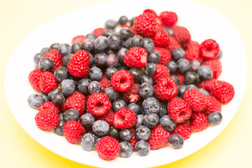 Forest berries, raspberries, blueberries in a bowl on a yellow background from above