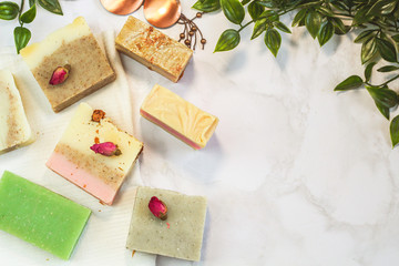 handmade craft soap different colors with herbs, flower petals. mint and rosebuds on a marble surface with green liana leaves on the side of flat lay. Spa treatment kit, body care with free space