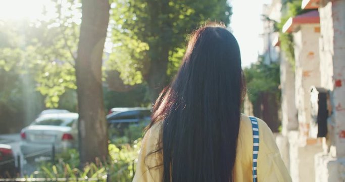 Back of lovely brunette long-haired young woman walking under sunshine in beautiful green residential street outdoors.