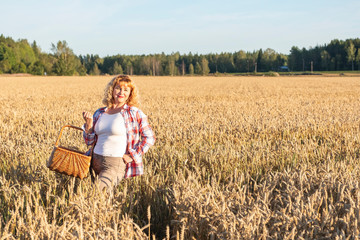 Naklejka na ściany i meble A red-haired girl stands in a field with ears of corn. Rural landscape. Blond European girl with wavy hair. Farmer with a wicker basket in his hands.