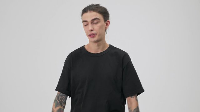 Annoyed young tattooed brunette man in black t-shirt disagree with someone and gesturing with hands while looking at the camera over gray background isolated