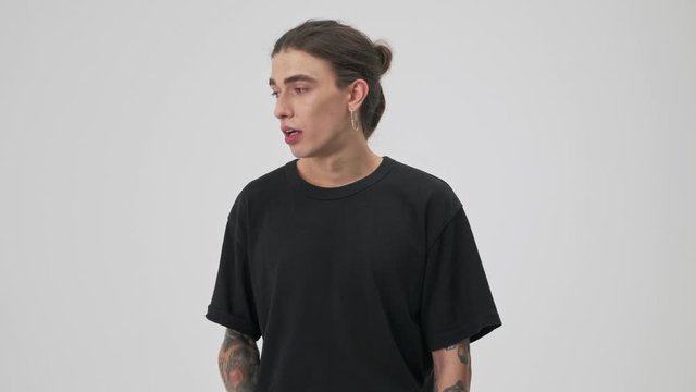 Displeased young tattooed brunette man in black t-shirt feeling nervous while looking at the camera over gray background isolated