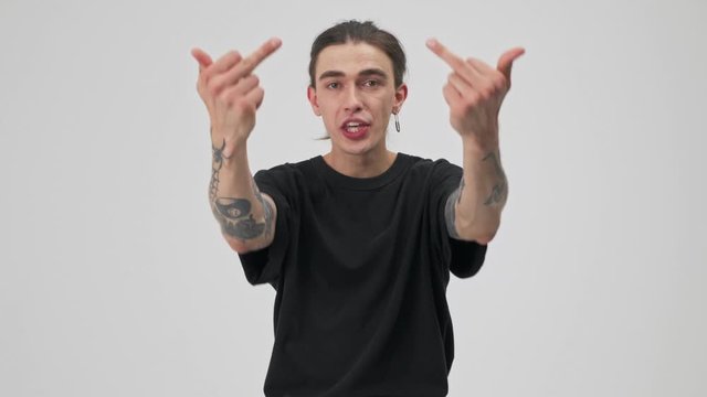 Angry young tattooed brunette man in black t-shirt arguing with someone and showing fuck you gesture with hands while looking at the camera over gray background isolated