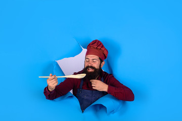Bearded man chef preparing to cook food. Handsome chef cook with kitchen utensils. Chief man in cook uniform with wooden spoon looking through paper. Bearded cook holds wooden kitchen cooking tools.