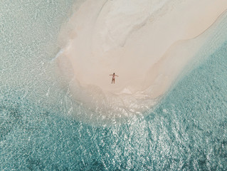 Aerial view of a sandbank in the Maldives sea - A young woman is lying on the white sand looking in the air with open arms
