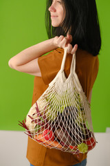 Young woman holds string bag mesh with vegetables and fruits on green background. Plastic free concept