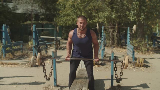 Brutal muscular bodybuilder doing heavy deadlift exercise at outdoor gym. Concentrated athletic fit man pumping up muscles with barbell, practicing upright row weight training during outdoor workout.