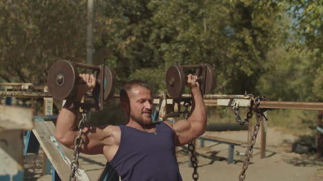 Concentrated bodybuilder practicing exrcise for chest with dumbbells while sitting on incline weight training bench at outdoors gym. Muscular fit man doing dumbbell chest press during outdoor workout