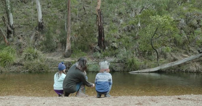 a mother is teaching her young children how to skip stones across a river. They are squatting on the river bank while the woman throws a stone that skips multiple times across the creek