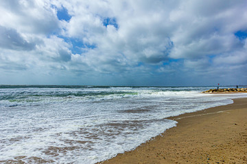 A view of a choppy and foamy sea along a sandy beach and cliff under a majestic blue sky and white clouds sky