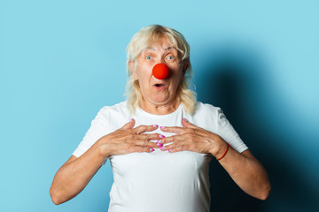 Old woman with a red clown nose makes hand gestures on a blue background. Red nose day concept,...