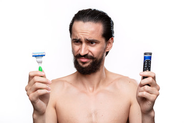 Bearded man with mustache holds electric razor and looks at the ols shaving stick standning bare isolated over white background. Morning treatment and shaving. Time to trim your beard.