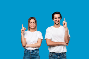 Happy couple in white t-shirts and jeans pointing up with fingers standing isolated over blue background. Copy space for product advertisement.