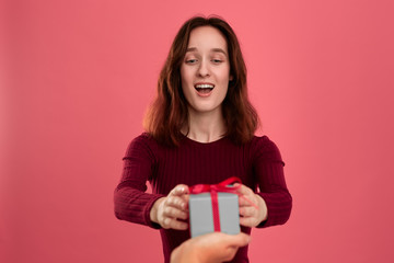 Happy pretty brunette girl surprised getting  present box with a ribbon from another person standing isolated on a dark pink background and smiling at the camera. Celebrating special event.