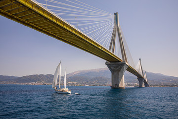 Sailboat passes under the Rio Antirrio Bridge or Charilaos Trikoupis Bridge, photo taken from the boat in summer sunny day.