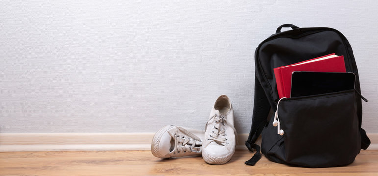 Old white sneakers, book and tablet with earphones in backpack on wooden background. School and student travel concept