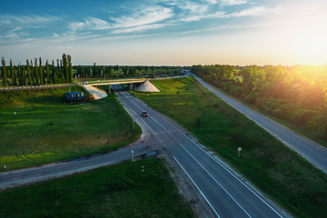 Road junction of speedy country highway route among green fields and trees with fast moving cars and trucks on background of beautiful dramatic sunset in warm summer evening