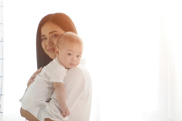 Beautiful Asian mother cradling her newborn baby on shoulder with happy face of being motherhood at home with white curtains background. Warm moment of mother and child, love and lifestyle concept