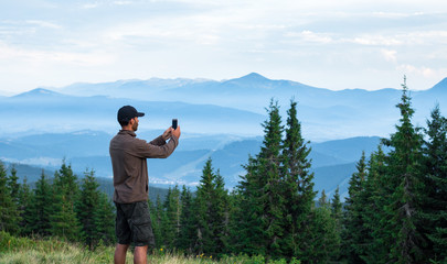 hiker man in cap taking photos of mountain range covered with blue fog in carpathian mountains