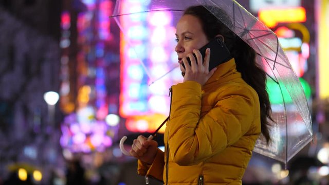 Tourist woman stay under umbrella at night city street, talk by phone. Bright blurred city lights on background. Lady wear warm jacket, while travelling around China at early spring season